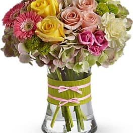Round mixed bouquet of pink gerberas, roses, and hydrangea in a clear vase wrapped with green and pink ribbon.