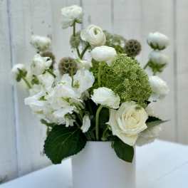 White roses and ranunculus with green accent blooms arranged in a white cylinder vase.