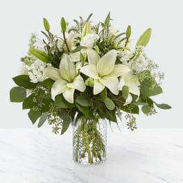 White lilies and mixed greenery arranged in a clear glass vase