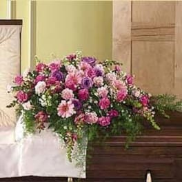 Pink and purple mixed flower casket spray with trailing greenery on a wooden casket.