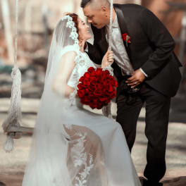 Bride holding a red rose bouquet while leaning toward groom on a swing