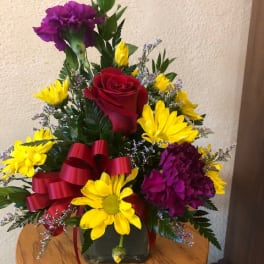 Red rose and yellow daisy arrangement with purple carnations in a glass vase