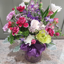 Colorful mixed bouquet of pink, purple, and white flowers in a purple vase with a ribbon and butterfly pick.