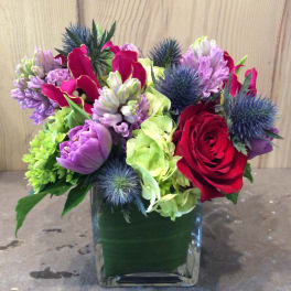 Bouquet of red roses, purple flowers, and blue thistles in a square glass vase