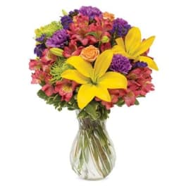 Mixed bouquet of yellow lilies, pink flowers, and purple blooms in a glass vase