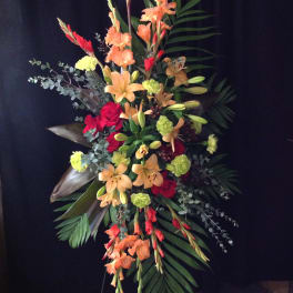 Tall standing spray with orange lilies, red roses, green carnations and gladiolus on a black backdrop