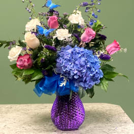 Mixed bouquet of blue hydrangea, pink roses, and white blooms in a purple glass vase
