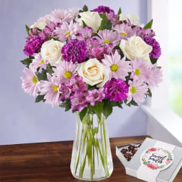Pink and white flowers in a clear glass vase beside a box of chocolates