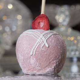 Pink candy-coated apple with white drizzle and a strawberry slice on top, set on a reflective surface.
