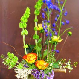 Tall mixed flower arrangement in a round vase with orange roses and blue blooms