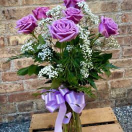 Lavender roses arranged in a glass vase with white filler flowers and a purple ribbon.