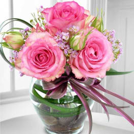 Small arrangement of pink roses and tiny lavender blooms in a round glass vase with curved green loops and a purple bow.