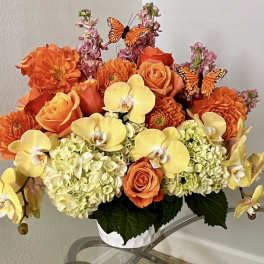 Orange and yellow floral arrangement with roses, orchids, and butterflies in a white container