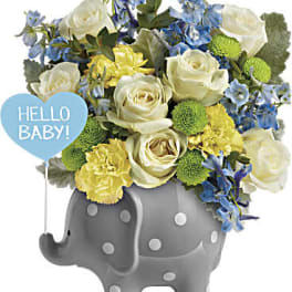 Bouquet of white roses, blue flowers, and yellow carnations in a gray elephant vase with a baby sign