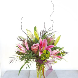 Pink lilies in a clear glass vase with greenery and a purple ribbon