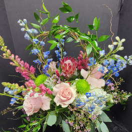 Pink roses and blue flowers arranged in a black container with a leafy arch