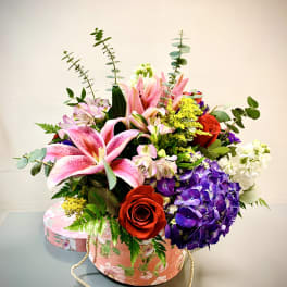 Mixed bouquet with pink lilies, red roses, purple hydrangea, and white blooms in a floral hatbox