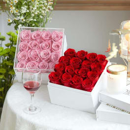 Two boxed rose arrangements in pink and red with a wine glass and candle nearby