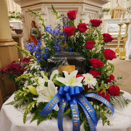 Large funeral floral arrangement with red roses, white lilies, and blue flowers