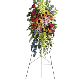 Tall standing floral spray with roses, lilies, sunflowers, and blue accents on an easel