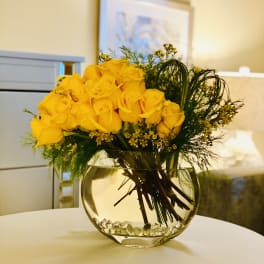 Yellow roses arranged in a round glass vase with greenery