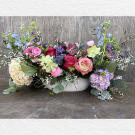 Low floral centerpiece with pink, purple, blue, and cream blooms in a white bowl.