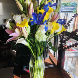 Tall bouquet of yellow calla lilies, blue irises, and pink lilies in a clear glass vase