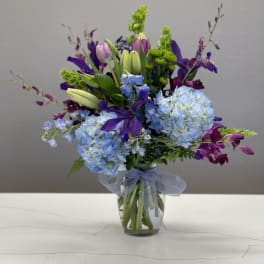 Mixed bouquet of blue hydrangeas, purple lilies, and green accents in a glass vase