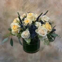 Cream roses arranged in a dark green glass vase with blue thistle accents