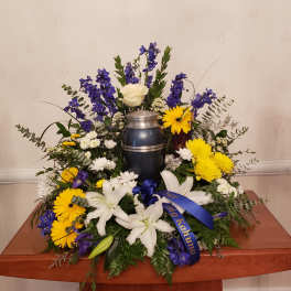 Sympathy floral arrangement with white lilies, yellow daisies, and blue flowers around an urn
