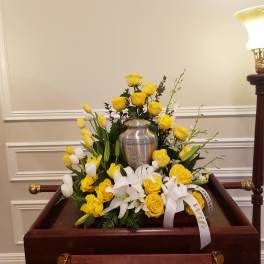 Yellow roses and white lilies arranged around a silver urn on a casket