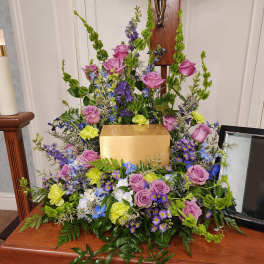 Large floral arrangement with pink roses, purple flowers, and green accents around a gold urn