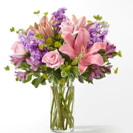 Pink lilies and roses in a clear glass vase with purple flowers
