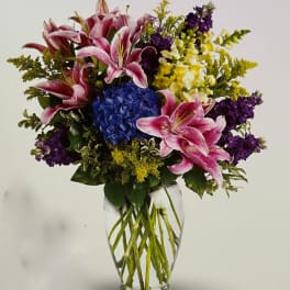 Bouquet of pink lilies, blue hydrangea, and purple flowers in a clear glass vase
