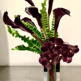 Tall arrangement of dark calla lilies and purple orchids in a glass vase