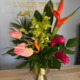 Tropical arrangement with green orchids, pink blooms, and orange heliconia in a gold pedestal vase