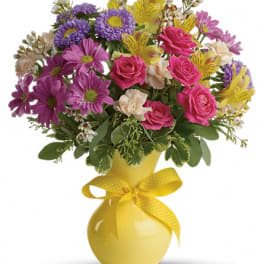 Mixed bouquet of pink, purple, and yellow flowers in a yellow vase with a ribbon