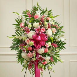 Tall standing spray of pink lilies, roses, carnations, and white mums with a large pink bow on an easel.