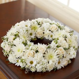 White and cream floral wreath of roses, daisies, and mums arranged in a ring on a wooden surface.