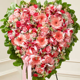 Heart-shaped standing spray of pink lilies, roses, and gerbera daisies with a green border and pink ribbon.