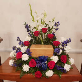 Symmetrical floral arrangement with red roses, white flowers, and blue accents around a wooden urn