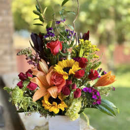 Mixed bouquet in a white square vase with roses, sunflowers, lilies, and purple blooms