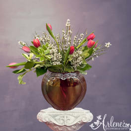 Pink tulips arranged in a heart-shaped glass vase with white filler flowers