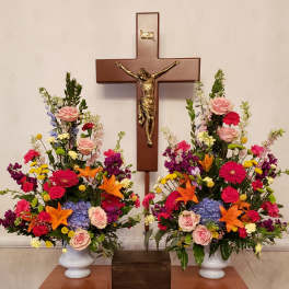 Two colorful floral arrangements flank a wooden crucifix on a table.