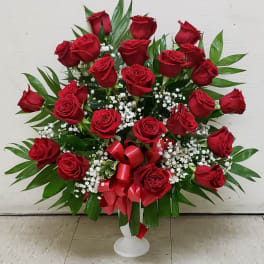 Red roses arranged in a white pedestal vase with baby's breath and a red ribbon