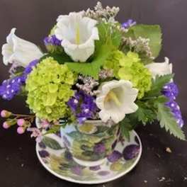 Compact arrangement of white and green flowers with purple accents in a patterned teacup and saucer