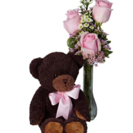Pink roses in a glass vase beside a teddy bear with a pink bow
