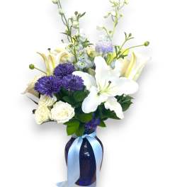 White lilies and purple flowers in a blue glass vase with a ribbon