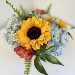 Compact bouquet with a large yellow sunflower, blue hydrangeas, and coral roses.