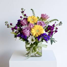 Mixed bouquet of purple roses, yellow gerberas and white daisies in a square glass vase with striped ribbon